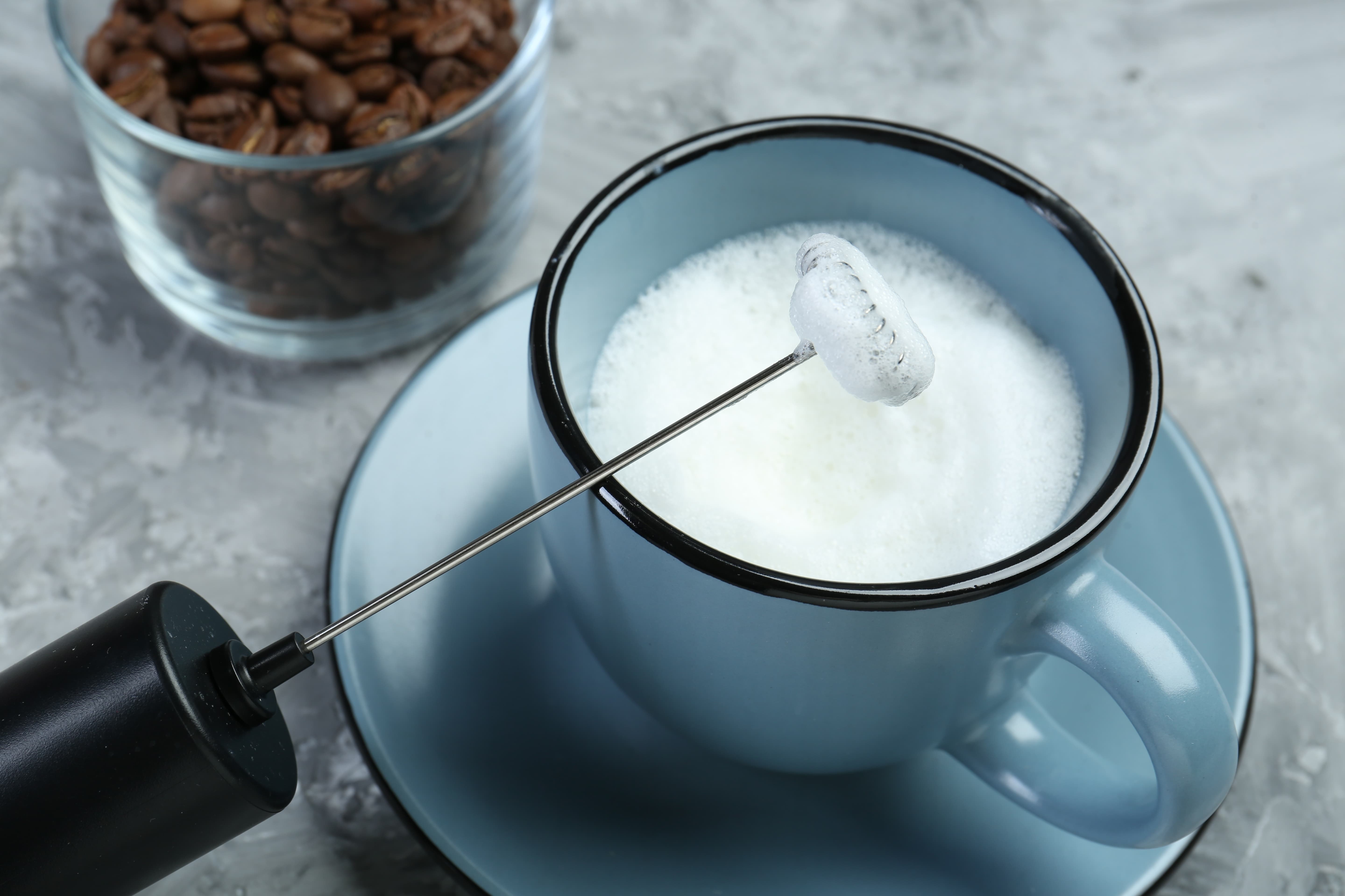 Milchaufschäumer erzeugt cremigen Milchschaum in einer Tasse, daneben Kaffeebohnen im Glas.