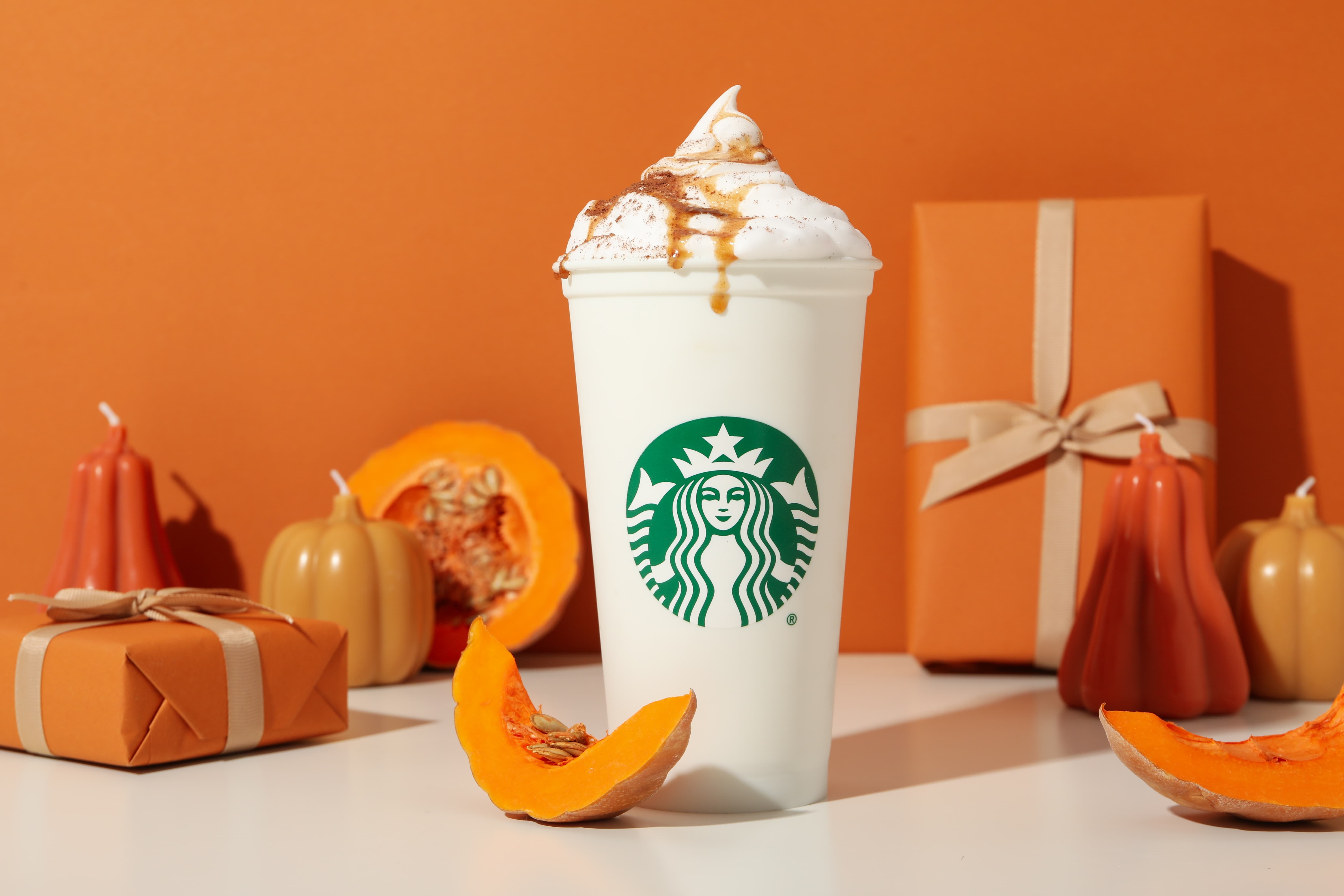 Starbucks Becher mit Pumpkin Spice Latte, Kürbis und Deko im Hintergrund.