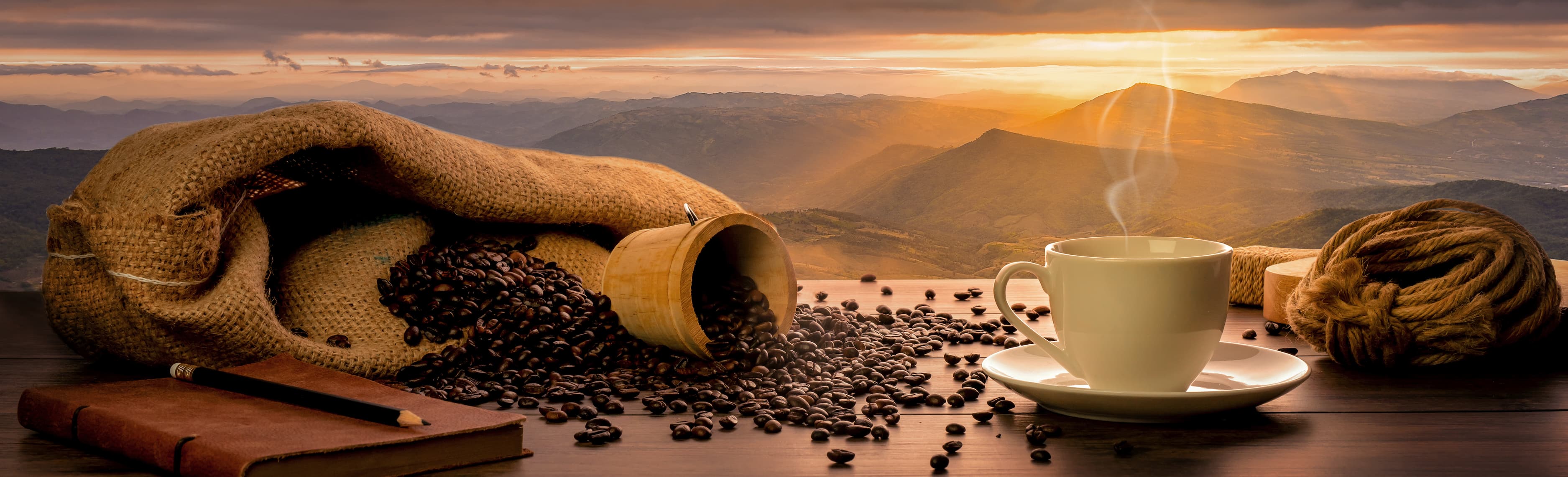Weiße Kaffeetasse mit Dampf neben Kaffeesack und Bohnen vor Sonnenaufgang über Berglandschaft.