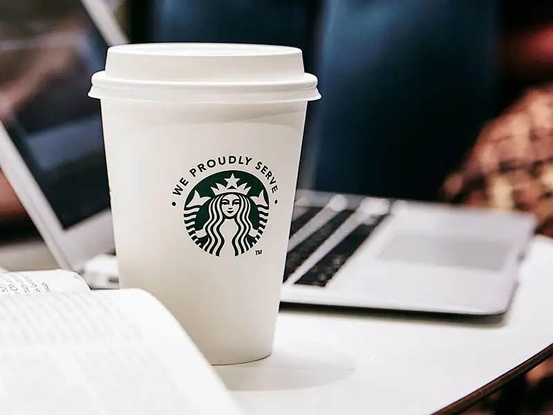 Starbucks Einwegbecher auf Tisch neben Laptop und Buch in gemuetlicher Umgebung.