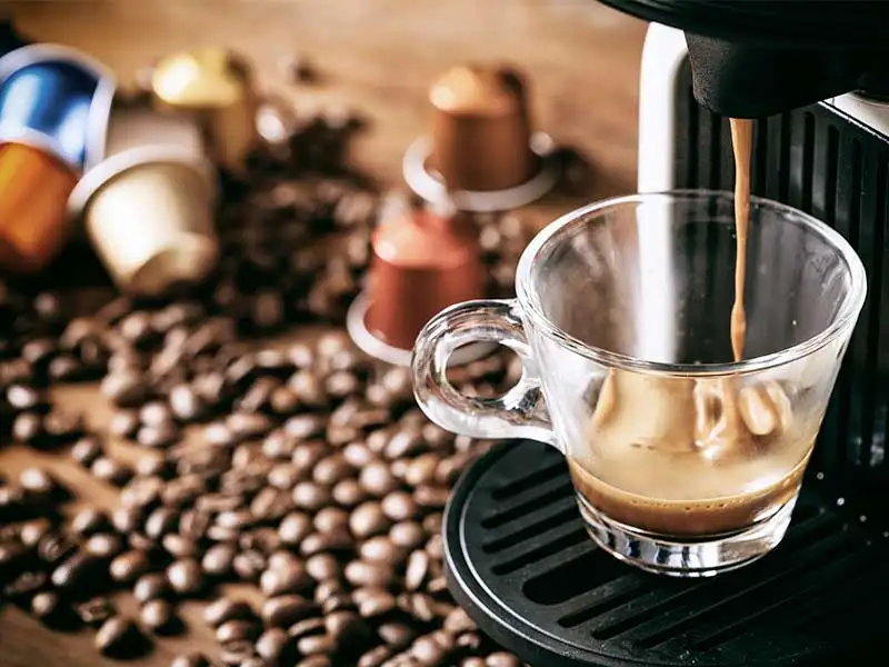 Espresso fließt aus der Kapselmaschine in eine Glastasse, im Hintergrund Kapseln und Bohnen.